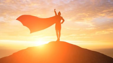 Stock photo of a female superhero standing atop a mountain, fist to the sky, cape billowing in the wind. It is sunrise.