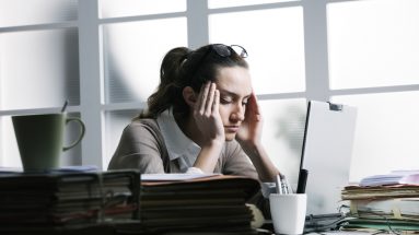 Stock photo of a woman sitting and working on her laptop surrounded by stacks of paper. She is frustrated and overwhelmed.