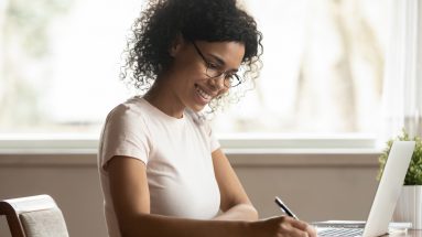 Stock photo of a woman in glasses sitting near computer holding pen writing